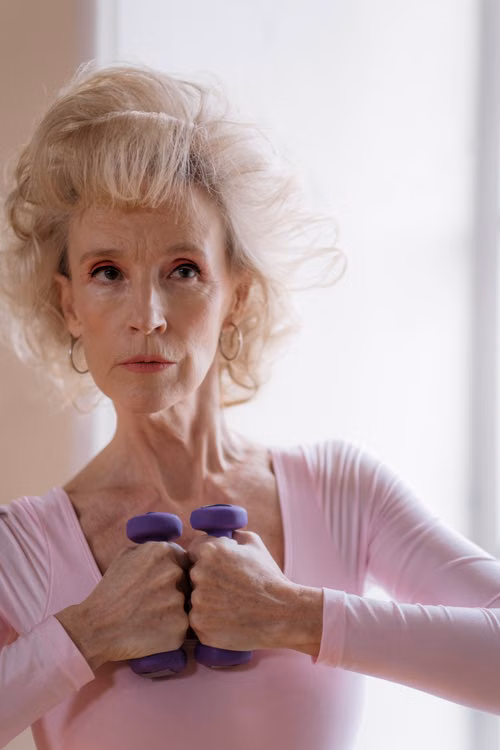 An older woman in pink holding a pair of purple dumbbells by a window.